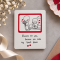 A square white greeting card with a cute cartoon illustration of a boy holding flowers and a girl sitting on a bench, surrounded by red hearts. Below the illustration, the card reads, 'Flowers for you, because you make my heart bloom' with two small red hearts.