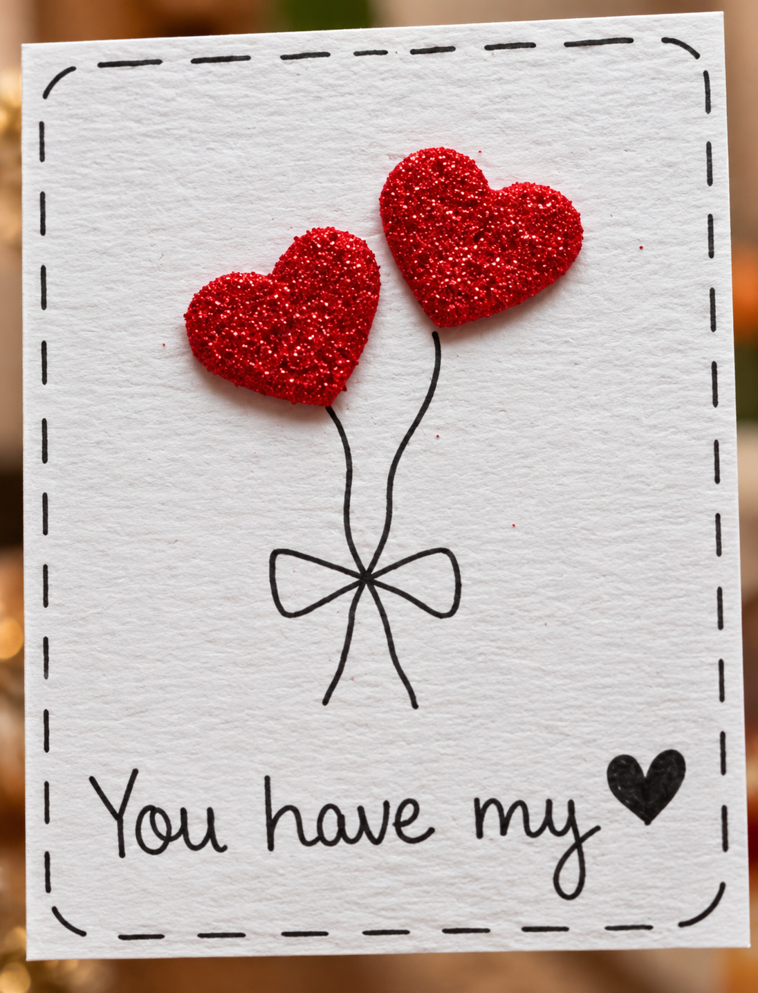 A square white handmade card with a stitched border, featuring two red glitter heart balloons tied with a black bow and the handwritten text 'You have my' with a small black heart. The background is softly blurred with warm bokeh.