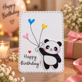 A handmade birthday card featuring a cute cartoon panda holding blue, yellow, and pink heart-shaped balloons, with 'Happy Birthday!' written on it. The card is displayed on a wooden table with a blurred background of a lit candle, gift box, and flowers.