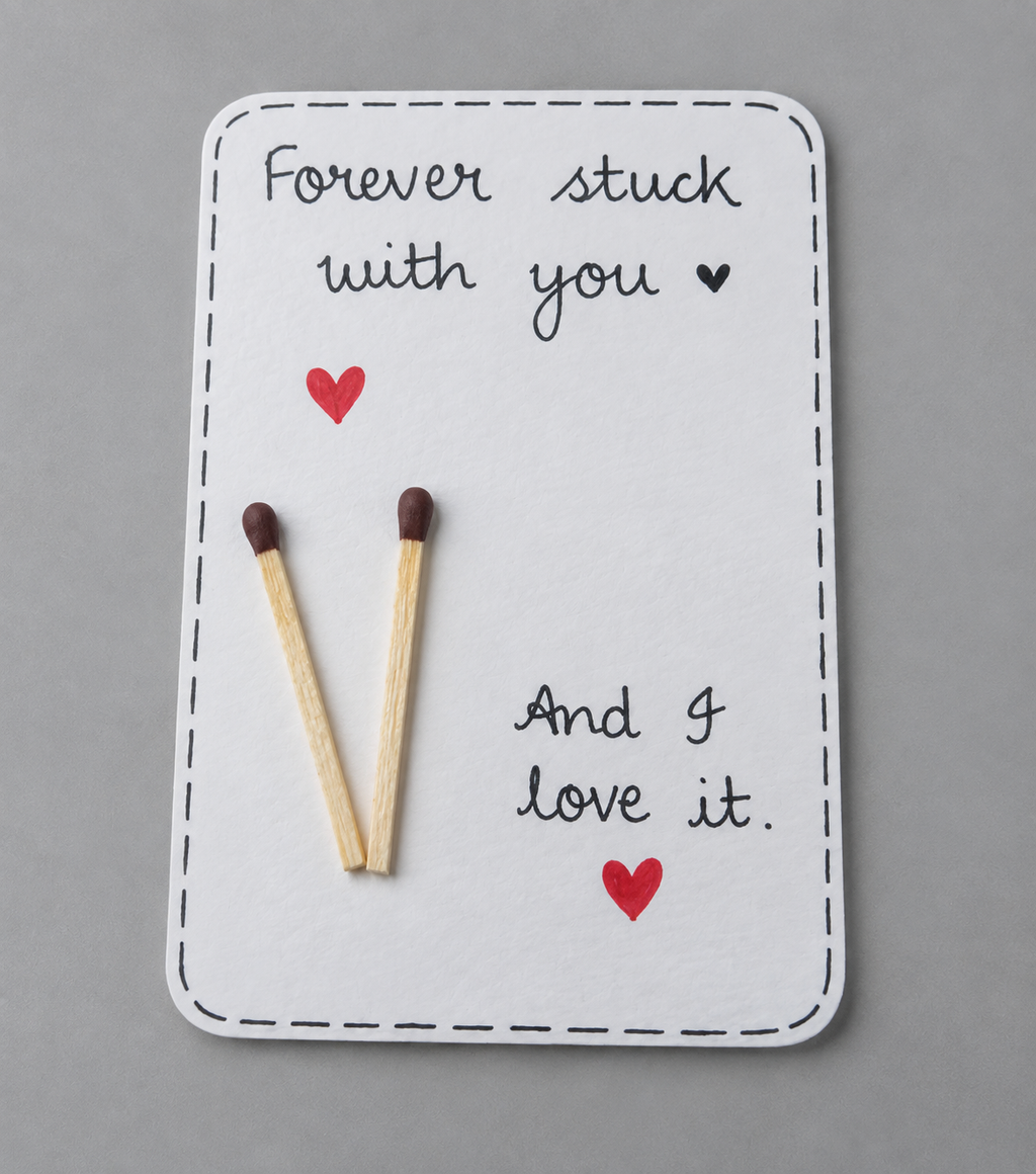 A square white greeting card with a dashed border, featuring the handwritten message 'Forever stuck with you' above two wooden matchsticks, and 'And I love it.' below, accented with two small red hearts, photographed on a smooth gray surface.