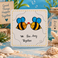 A square white card with rounded corners featuring a cartoon bee illustration and the phrase 'We Bee-Long Together.' The card is styled on a beach-themed surface with sand, shells, greenery, and a small jar tag reading 'You & Me.