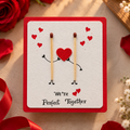 A square greeting card with a red border and a white center, featuring two matchsticks holding a red heart with the words 'We're Perfect Together' below. The card is styled on a beige surface with red roses, a red ribbon, and small white flowers.