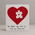A square white card with a large red heart, a white puzzle-piece cutout in the center, and black handwritten text that reads, 'You lightup my world by fixing my missing piece.' The card is photographed against a soft gray background.
