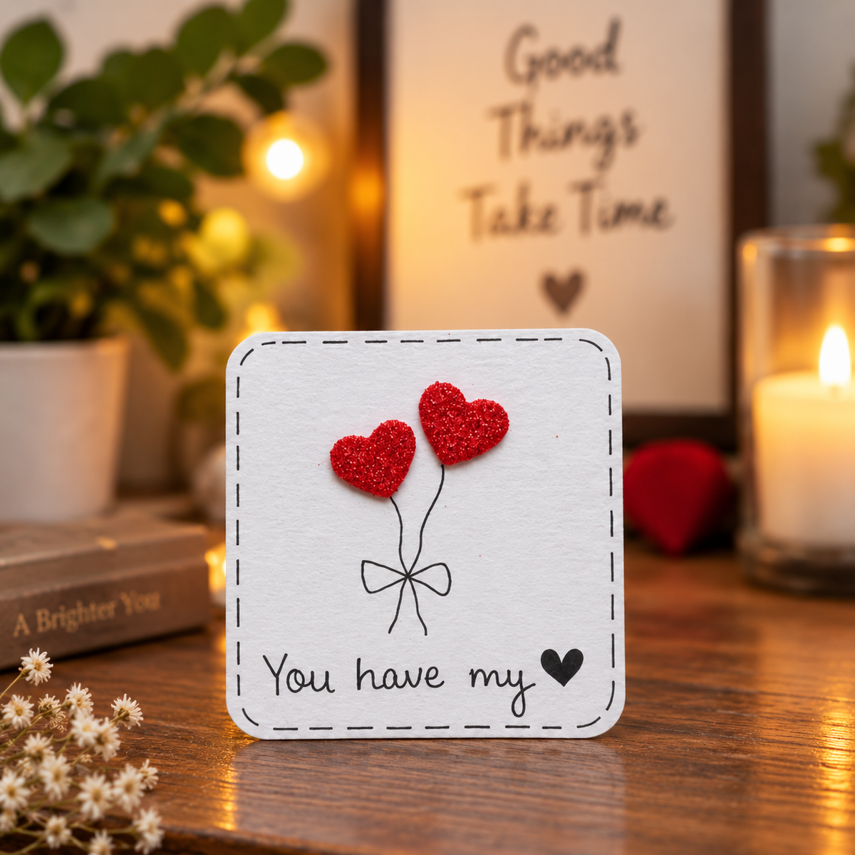 A square white card with a stitched border design, featuring two red glitter heart balloons and the text 'You have my heart' on a wooden table. The background is warmly lit with a blurred sign reading 'Good Things Take Time', a lit candle, and a potted plant.
