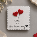 A square white card with rounded corners featuring a black dashed border, a simple line drawing of two red glitter hearts tied with a bow, and the phrase 'You have my' with a black heart. The card is photographed on a soft gray surface with small decorative props around it.