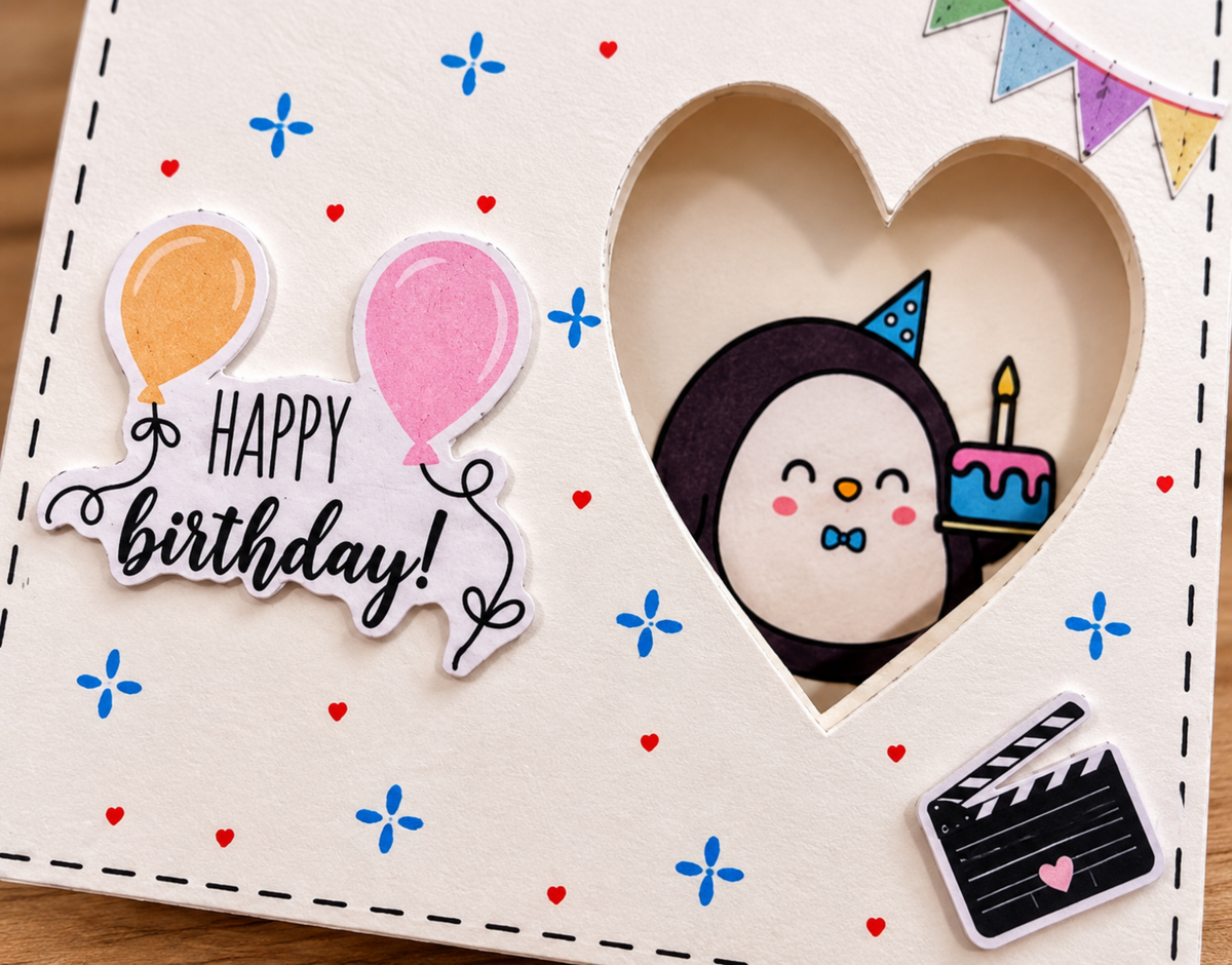 A close-up of a handmade birthday card featuring a heart-shaped cutout with a penguin wearing a party hat and holding a birthday cake. The card is decorated with blue and red starburst patterns, a 'Happy birthday!' banner with balloons, and a clapperboard sticker.