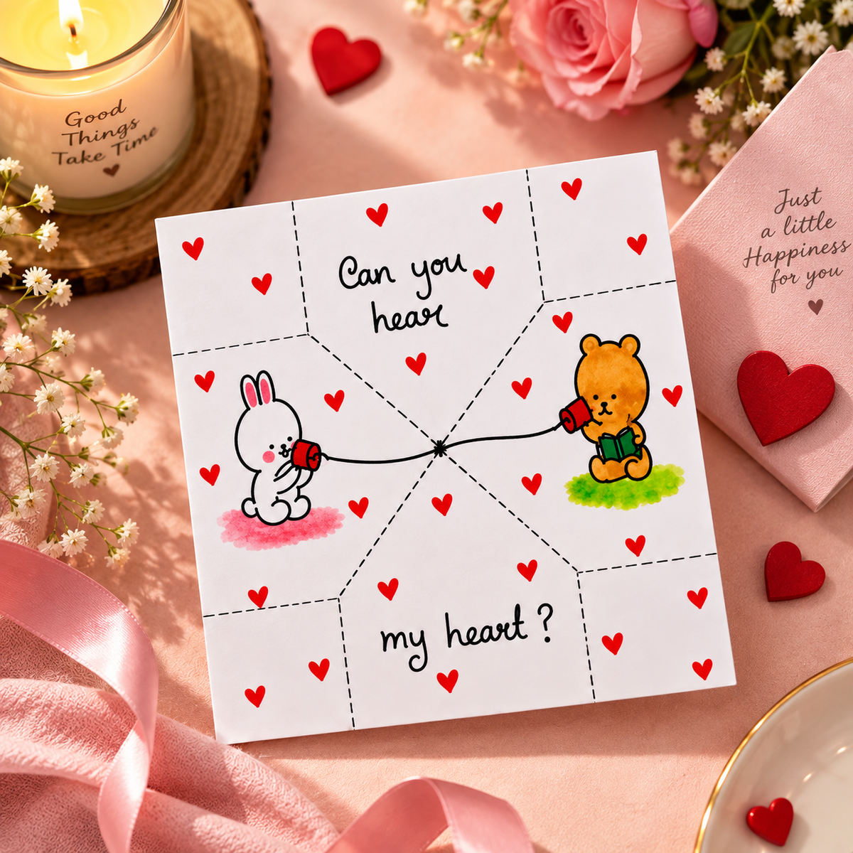 A square white greeting card with a heart-themed illustration, placed on a pink surface with flowers, ribbon, a candle, and a pink envelope around it. The card reads 'Can you heart my heart?' with a bunny holding a red heart on the left and a bear holding a green heart on the right.