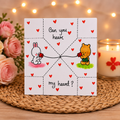 A square white greeting card with a heart-themed cartoon design, placed on a woven placemat with pink roses and a lit candle in the background. The card reads 'Can you heart my heart?' with illustrations of a white bunny and a yellow bear holding red hearts.