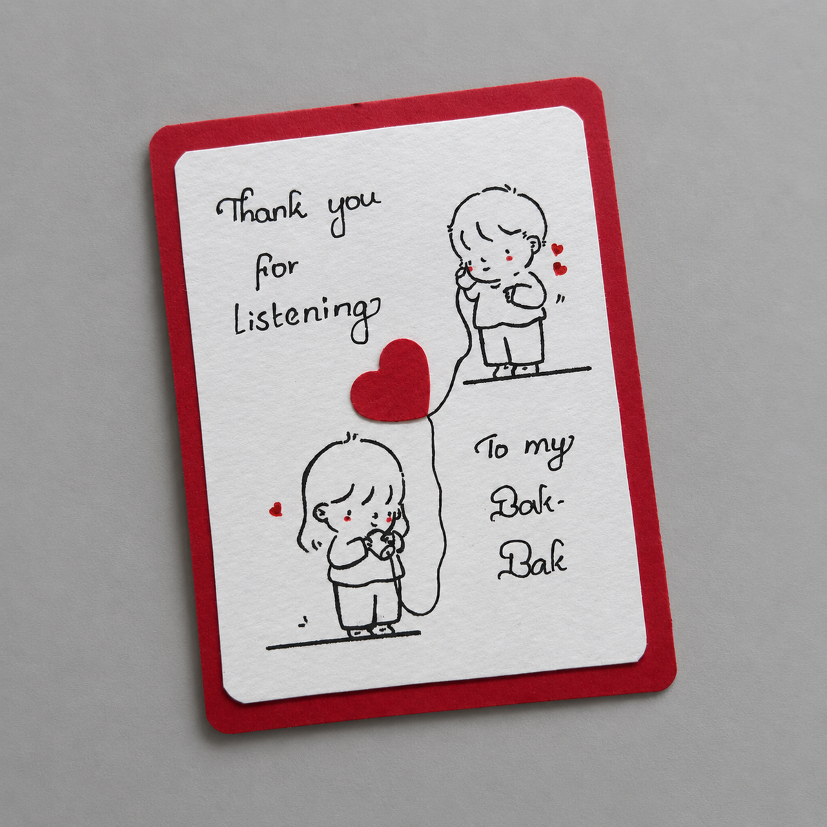 A handmade greeting card with a red border and white center, featuring black line drawings of children holding red heart-shaped balloons and handwritten text that reads 'Thank you for Listening' and 'To my Bak-Bak'. The card is photographed on a neutral gray background.