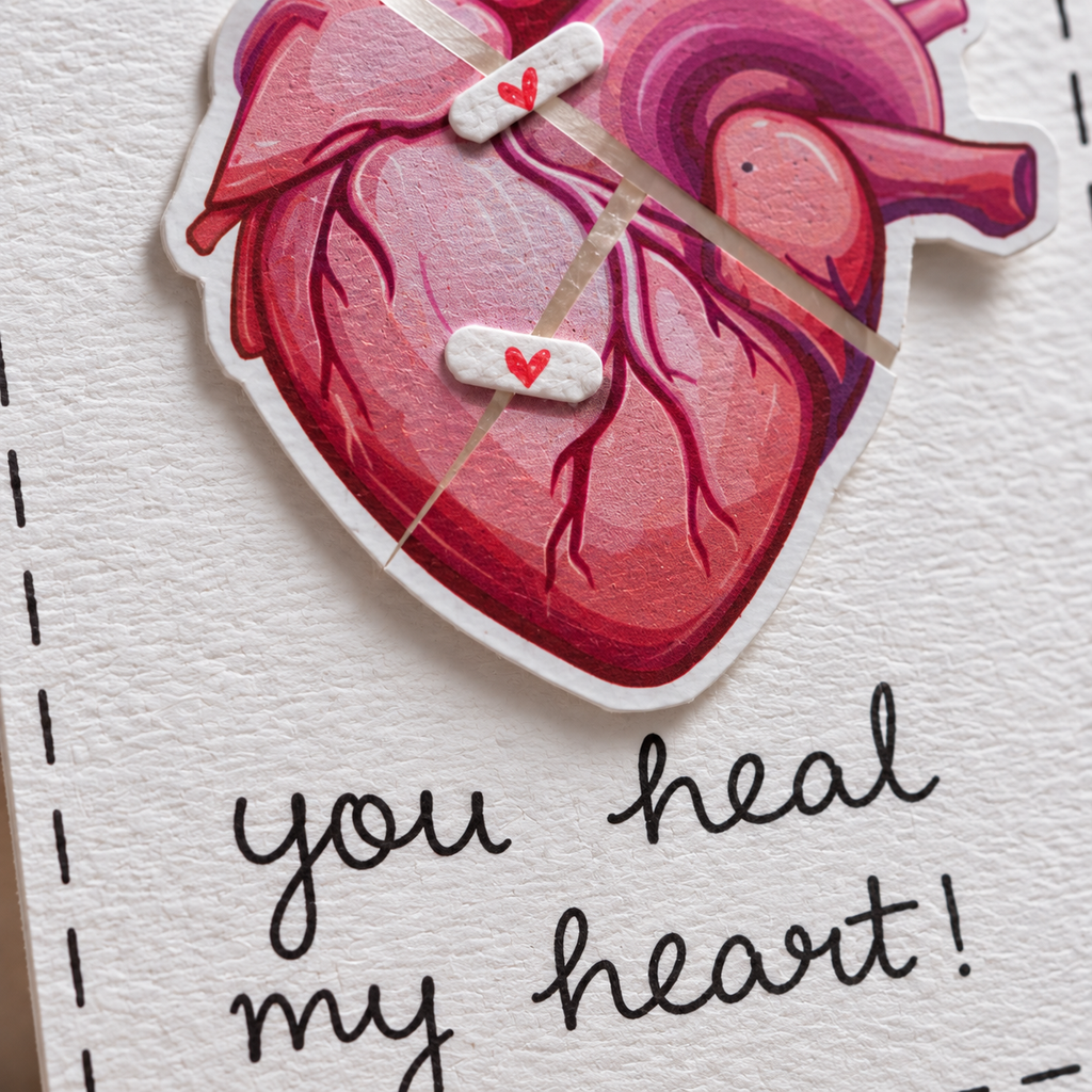 A close-up of a handmade greeting card featuring a die-cut illustration of a pink and red anatomical heart with two white healing bandages across it, each with a small red heart. Below the heart, the card reads 'you heal my heart!' in black script on textured white cardstock.