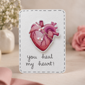 A square white greeting card with a stitched border, featuring a pink anatomical heart clock face and the handwritten message 'you heal my heart!' in black script. The card is photographed on a soft beige surface with blurred pink flowers and a small white vase in the background.