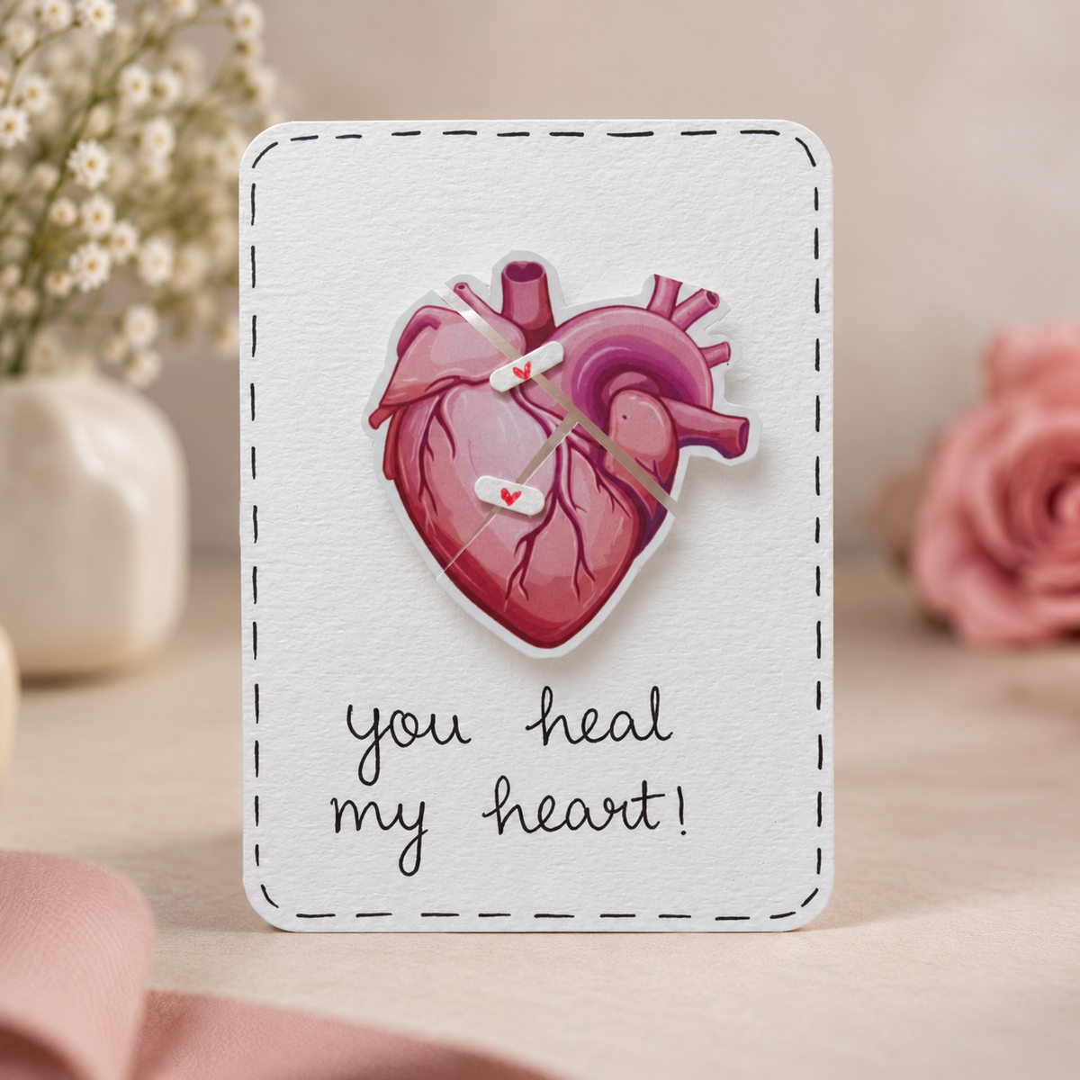 A square white greeting card with a stitched border, featuring a pink anatomical heart clock face and the handwritten message 'you heal my heart!' in black script. The card is photographed on a soft beige surface with blurred pink flowers and a small white vase in the background.