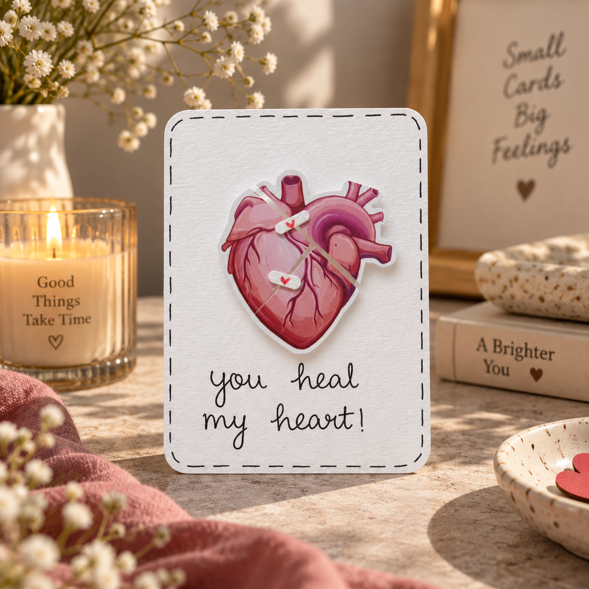 A square white greeting card with a stitched border, featuring a pink anatomical heart clock and the words 'you heal my heart!' in black script. The card is styled on a tabletop with a lit candle, dried flowers, a framed sign reading 'Small Cards Big Feelings,' and a book.